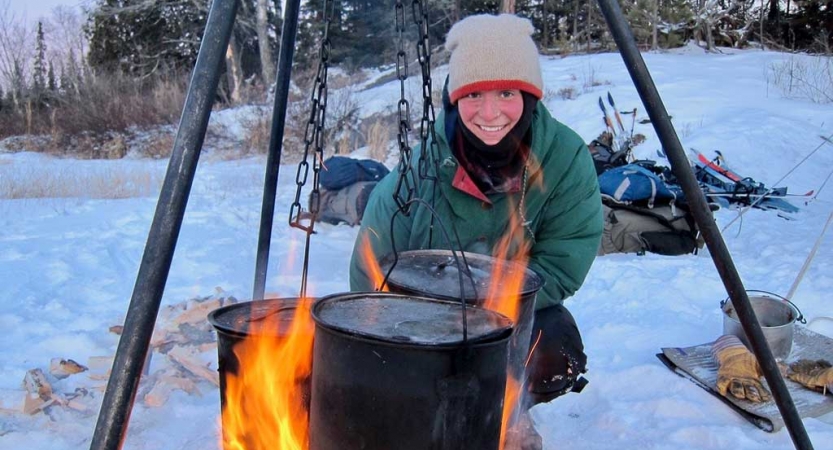 A person smiles while kneeling beside a tripod holding pots over a campfire in a snowy landscape. 